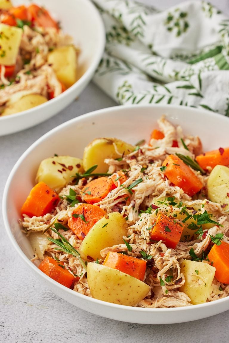 Slow cooker honey chicken with carrots and potatoes in a white bowl, featuring shredded chicken, tender baby potatoes, and honey-glazed carrots with fresh herbs